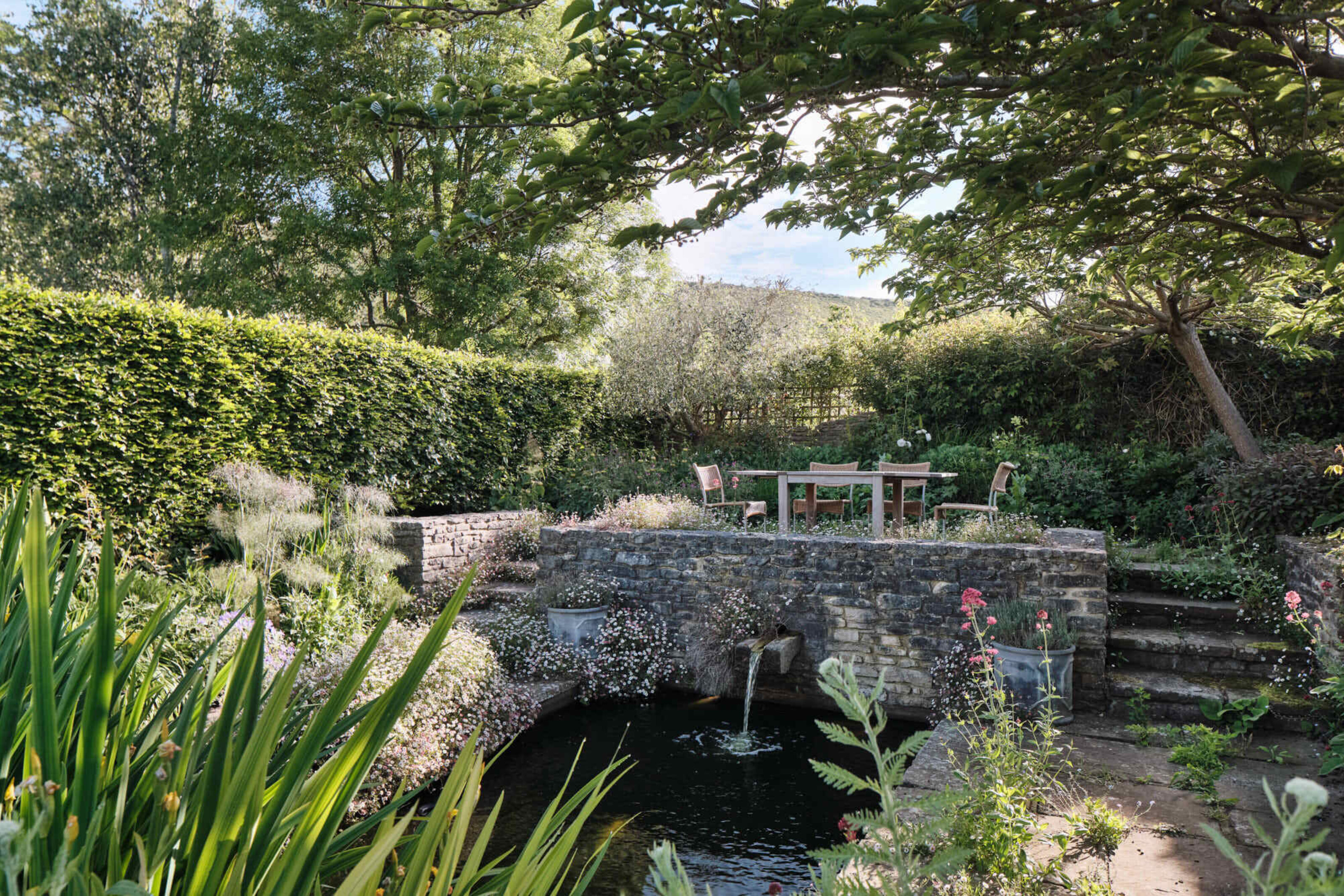 Landscaped garden terrace overlooking a pond at Corfe Castle House, surrounded by naturalistic planting, stone walls and mature trees.