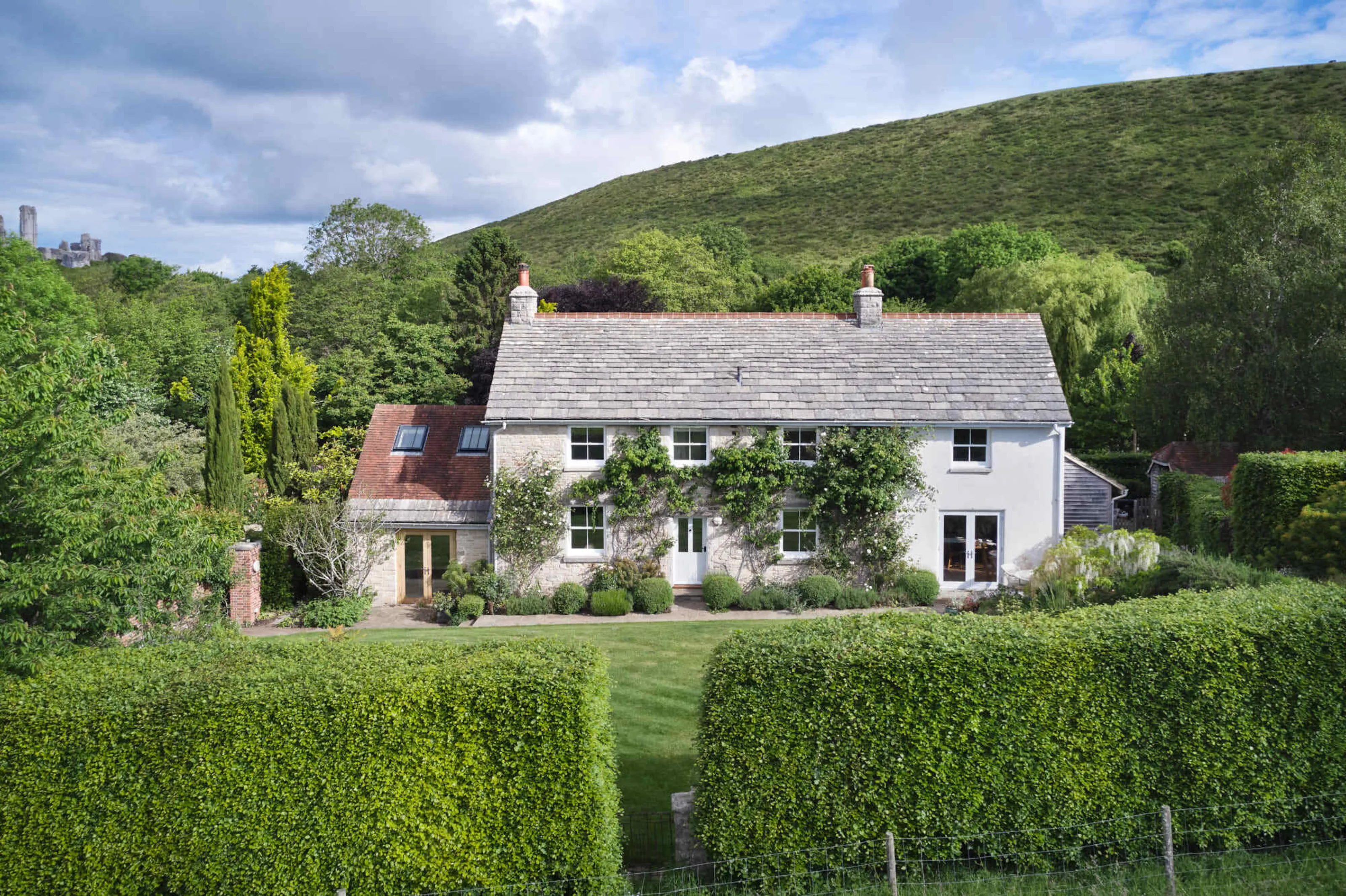 Front elevation of Corfe Castle House, showing the restored cottage set behind clipped hedges and surrounded by mature planting at the foot of the Purbeck Hills.