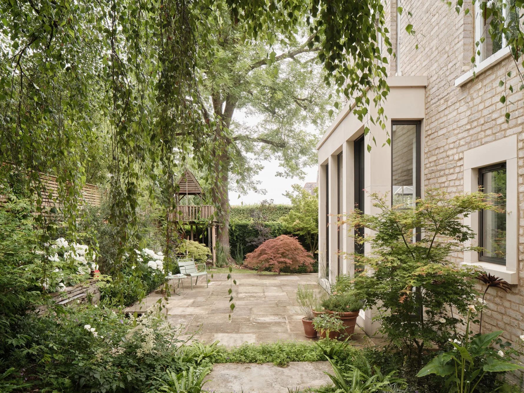 View across a mature garden showing the rear extension, stone terrace and established trees surrounding the family home.