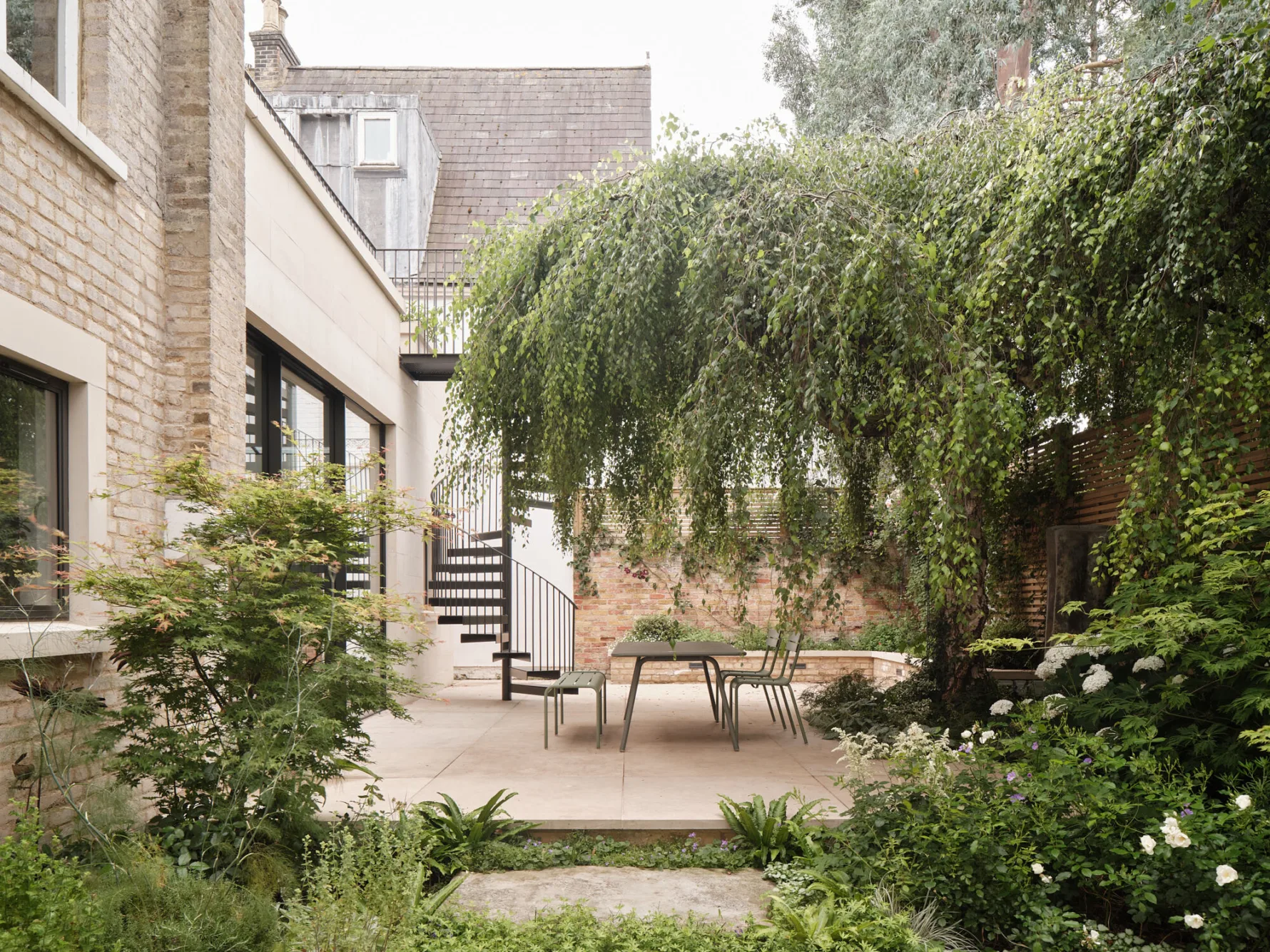 View across a mature garden showing the rear extension, stone terrace and established trees surrounding the family home.