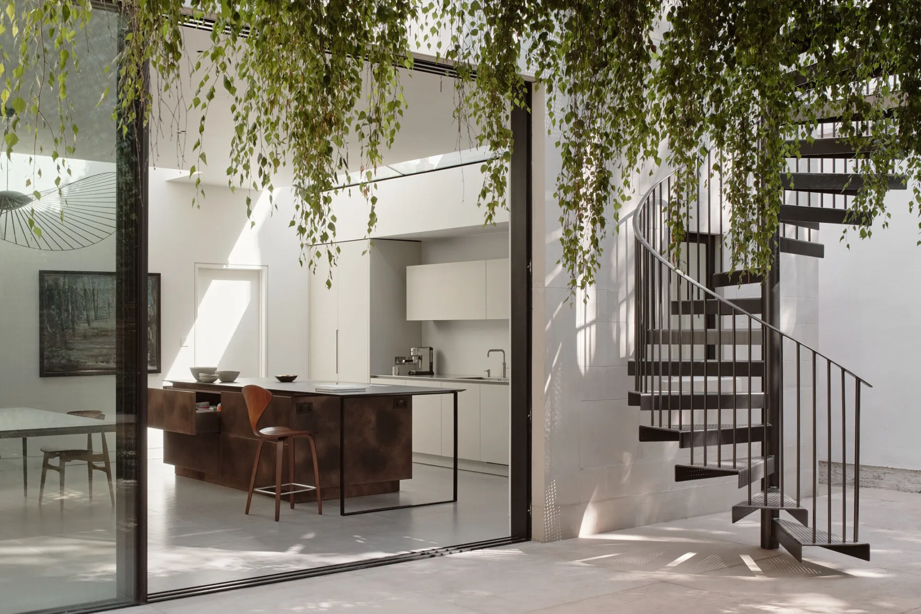 View from the courtyard into the contemporary kitchen extension, featuring full-height glazing, a sculptural staircase and a metal-clad kitchen island.