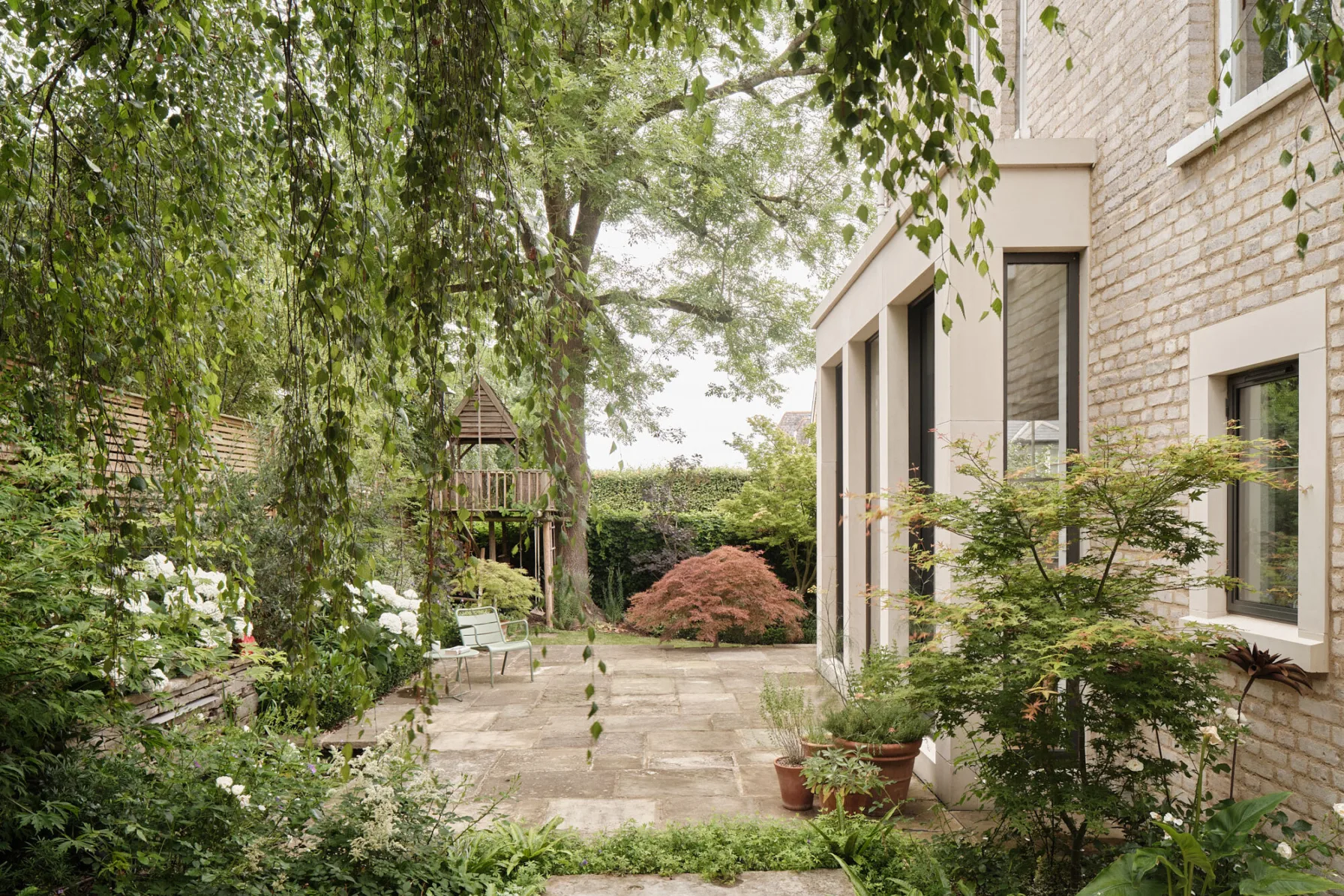 View across a mature garden showing the rear extension, stone terrace and established trees surrounding the family home.