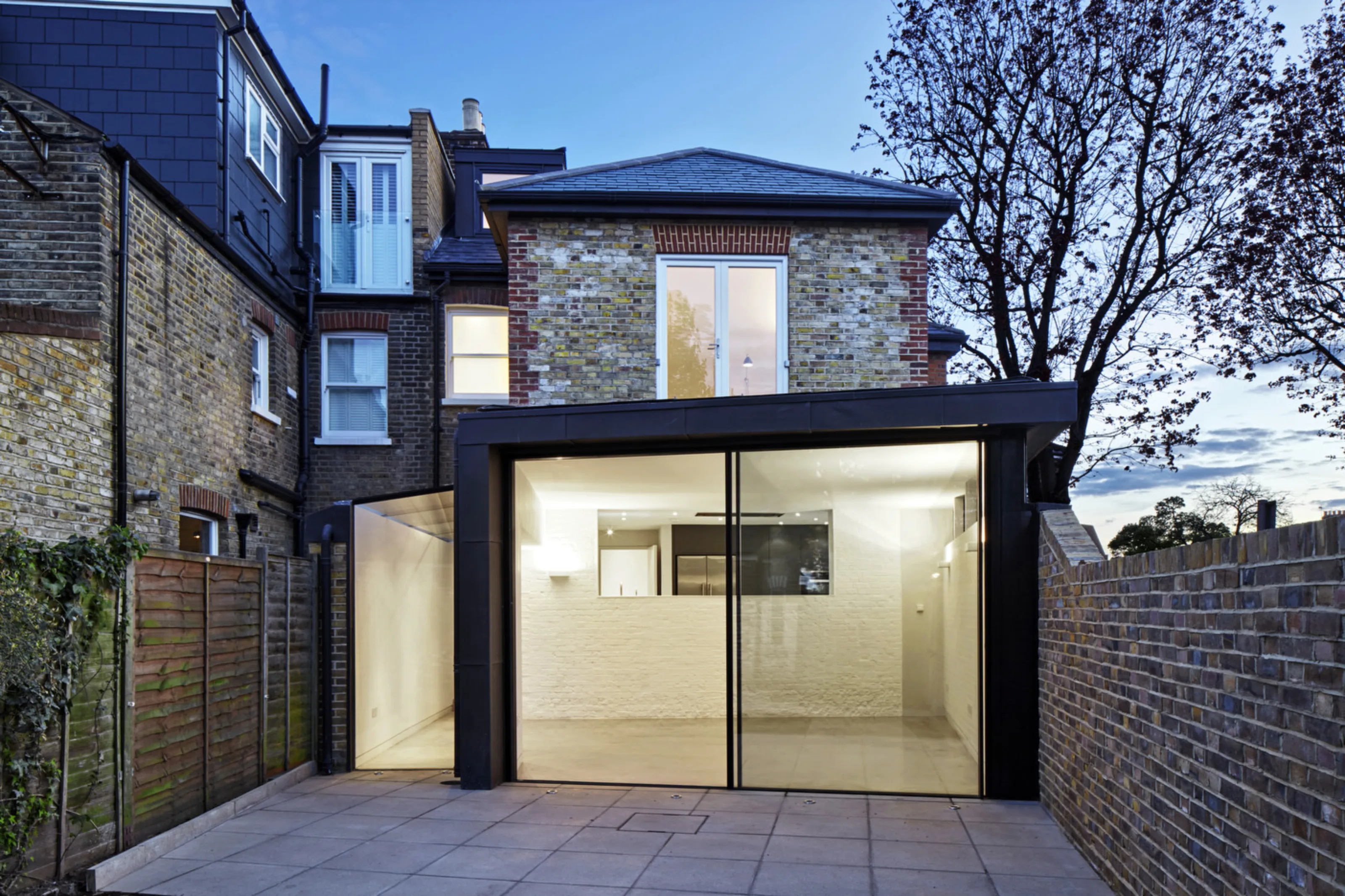 Twickenham House contemporary rear extension with zinc cladding, large sliding doors and garden terrace at twilight.